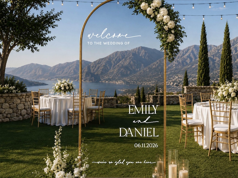 May include: A tall, arched gold metal wedding welcome sign featuring white text that reads: welcome TO THE WEDDING OF EMILY and DANIEL 06.11.2026 we're so glad you are here. The sign is decorated with a cluster of white flowers and greenery at the top.