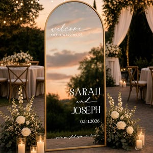 May include: An arched floor mirror with a thin gold frame featuring white printed text that reads: welcome TO THE WEDDING OF SARAH and JOSEPH 03.11.2026 we're so glad you are here. The mirror reflects a sunset sky and outdoor event space.