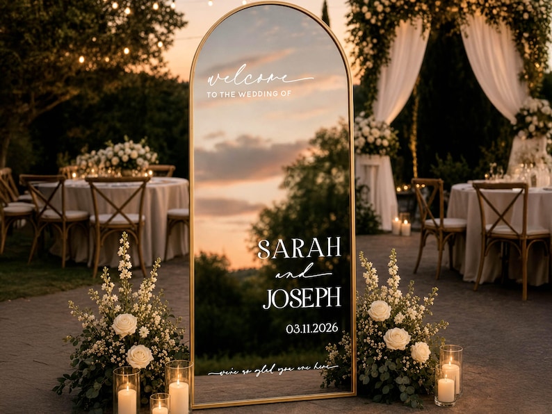 May include: An arched floor mirror with a thin gold frame featuring white printed text that reads: welcome TO THE WEDDING OF SARAH and JOSEPH 03.11.2026 we're so glad you are here. The mirror reflects a sunset sky and outdoor event space.