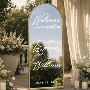 May include: An arched floor mirror with a gold frame featuring white text that reads Welcome to our wedding, Emma + William, and June 12, 2026. The reflective surface displays a clear view of the surrounding outdoor environment and trees.