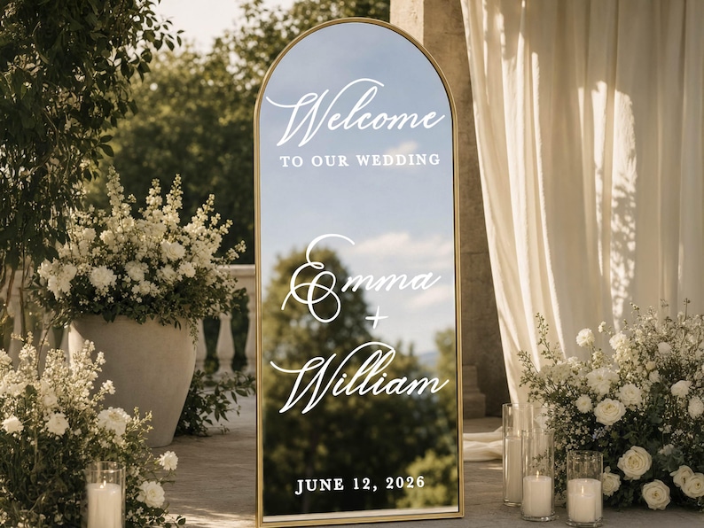 May include: An arched floor mirror with a gold frame featuring white text that reads Welcome to our wedding, Emma + William, and June 12, 2026. The reflective surface displays a clear view of the surrounding outdoor environment and trees.