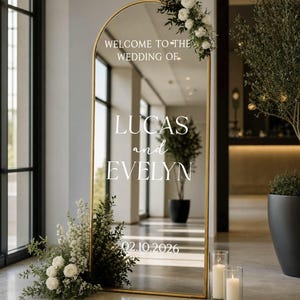 May include: An arched floor mirror with a gold frame featuring white text that reads WELCOME TO THE WEDDING OF LUCAS and EVELYN 02.10.2026. The mirror is decorated with white flowers and greenery at the top right and base, reflecting the surrounding interior space.