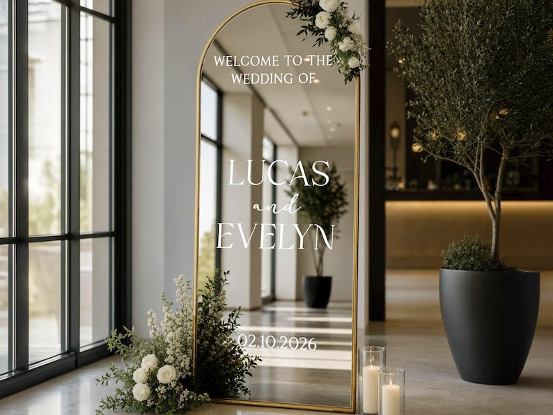 May include: An arched floor mirror with a gold frame featuring white text that reads WELCOME TO THE WEDDING OF LUCAS and EVELYN 02.10.2026. The mirror is decorated with white flowers and greenery at the top right and base, reflecting the surrounding interior space.