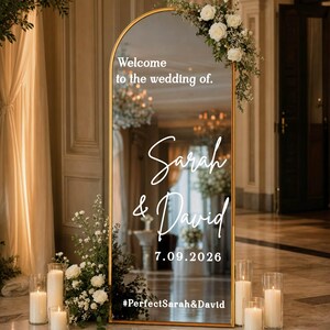 May include: An arched floor mirror with a gold frame featuring white text that reads Welcome to the wedding of, Sarah & David, 7.09.2026, and #PerfectSarah&David. The mirror is decorated with white floral arrangements and surrounded by glass pillar candles.