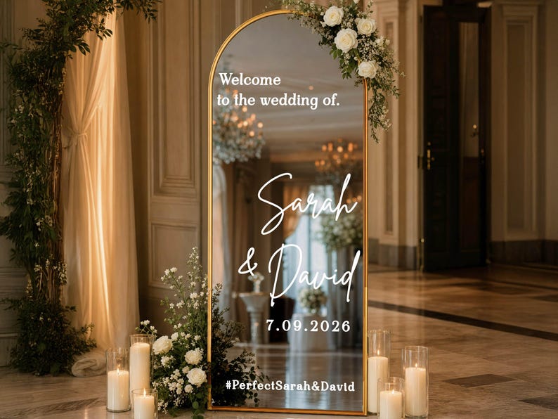 May include: An arched floor mirror with a gold frame featuring white text that reads Welcome to the wedding of, Sarah & David, 7.09.2026, and #PerfectSarah&David. The mirror is decorated with white floral arrangements and surrounded by glass pillar candles.