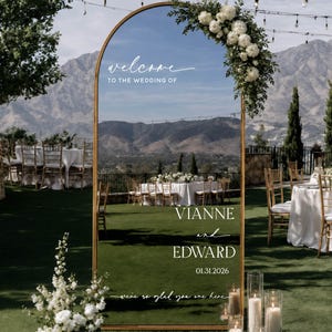 May include: An arched gold-framed mirror wedding sign featuring white text that reads: welcome TO THE WEDDING OF VIANNE and EDWARD 01.31.2026 we're so glad you are here. The mirror is decorated with a cluster of white flowers and greenery at the top.