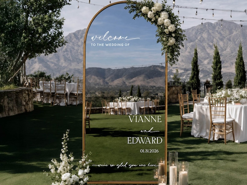 May include: An arched gold-framed mirror wedding sign featuring white text that reads: welcome TO THE WEDDING OF VIANNE and EDWARD 01.31.2026 we're so glad you are here. The mirror is decorated with a cluster of white flowers and greenery at the top.
