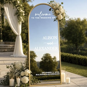 May include: An arched floor mirror with a thin gold frame featuring white text that reads: welcome TO THE WEDDING OF ALISON and ALEXANDER DECEMBER 3, 2026 #ALISON&ALEXWEDDING. The mirror reflects a landscape and is decorated with white flowers.