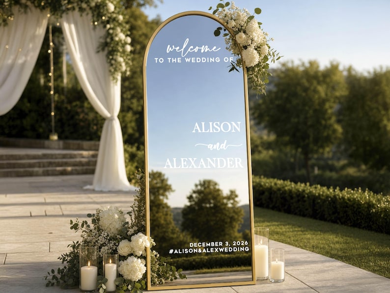 May include: An arched floor mirror with a thin gold frame featuring white text that reads: welcome TO THE WEDDING OF ALISON and ALEXANDER DECEMBER 3, 2026 #ALISON&ALEXWEDDING. The mirror reflects a landscape and is decorated with white flowers.