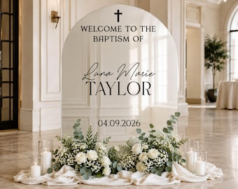 May include: An arched, reflective sign for a baptism featuring a black cross icon, the text "WELCOME TO THE BAPTISM OF", the name "Luna Marie TAYLOR", and the date "04.09.2026". The base is decorated with white roses, baby's breath, and eucalyptus.