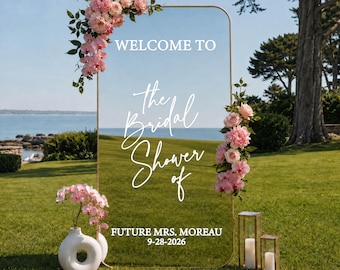 May include: A rectangular clear acrylic sign with a gold metal frame, decorated with pink floral arrangements. The text reads: WELCOME TO The Bridal Shower of FUTURE MRS. MOREAU 9-28-2026. A white ceramic vase with pink flowers sits nearby.
