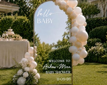 May include: An arched mirror sign with a gold frame, decorated with white balloons and flowers. The text reads: hello BABY, WELCOME TO Andrew + Mina BABY SHOWER, and 11.05.26. The mirror reflects the surrounding outdoor garden and lawn area.
