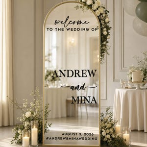 May include: An arched floor mirror with a gold frame featuring black text that reads: welcome TO THE WEDDING OF ANDREW and MINA AUGUST 3, 2026 #ANDREW&MINAWEDDING. The mirror is decorated with white flowers and greenery along the top right corner.