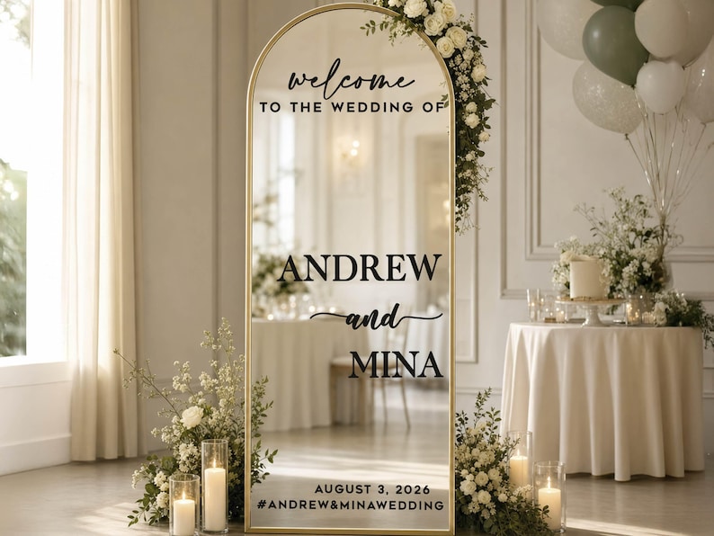 May include: An arched floor mirror with a gold frame featuring black text that reads: welcome TO THE WEDDING OF ANDREW and MINA AUGUST 3, 2026 #ANDREW&MINAWEDDING. The mirror is decorated with white flowers and greenery along the top right corner.