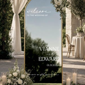 May include: An arched gold-framed mirror wedding welcome sign featuring white text that reads: welcome TO THE WEDDING OF VIANNE and EDWARD 01.31.2026 we're so glad you are here. The sign is decorated with white floral arrangements at the top and base.