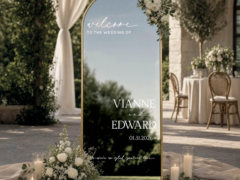 May include: An arched gold-framed mirror wedding welcome sign featuring white text that reads: welcome TO THE WEDDING OF VIANNE and EDWARD 01.31.2026 we're so glad you are here. The sign is decorated with white floral arrangements at the top and base.