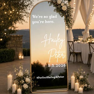 May include: An arched floor mirror with a thin gold frame featuring white text that reads We're so glad you're here, Hailey, Peter, 5.3.2026, and #PerfectHailey&Peter. The mirror is decorated with white floral arrangements and pillar candles.