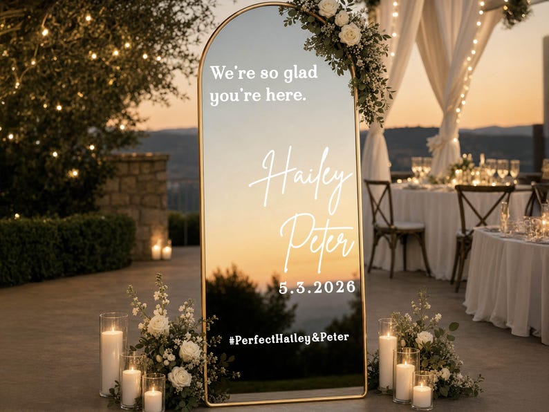 May include: An arched floor mirror with a thin gold frame featuring white text that reads We're so glad you're here, Hailey, Peter, 5.3.2026, and #PerfectHailey&Peter. The mirror is decorated with white floral arrangements and pillar candles.