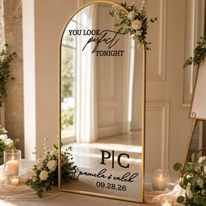May include: An arched floor mirror with a thin gold frame, decorated with white floral arrangements. The glass features black text reading "YOU LOOK perfect TONIGHT", the initials "P|C", the names "pamela & caleb", and the date "09.28.26".