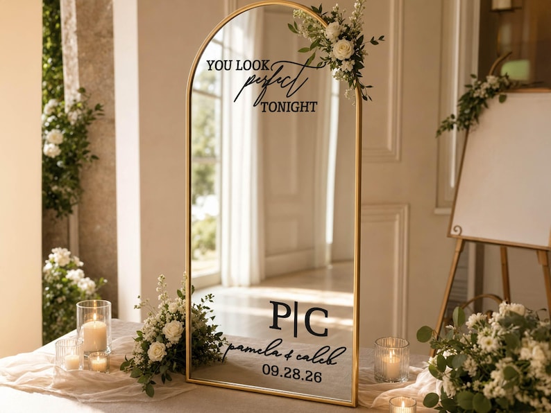 May include: An arched floor mirror with a thin gold frame, decorated with white floral arrangements. The glass features black text reading "YOU LOOK perfect TONIGHT", the initials "P|C", the names "pamela & caleb", and the date "09.28.26".