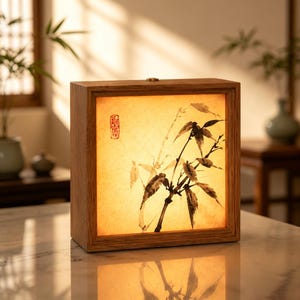 May include: A square wooden lamp with a light-colored frame and a translucent panel featuring a black ink-style illustration of bamboo stalks and leaves. A small red rectangular seal is visible on the left side of the illuminated panel.