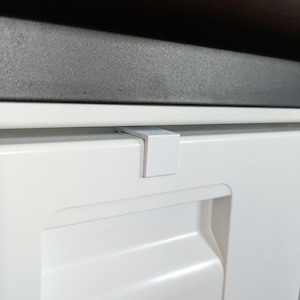 Peut inclure: Un petit clip en plastique blanc en forme de L fixé sur le bord supérieur d'une porte de placard blanche. Le clip présente une surface texturée et repose à plat contre l'avant du placard, qui comporte un panneau rectangulaire encastré.
