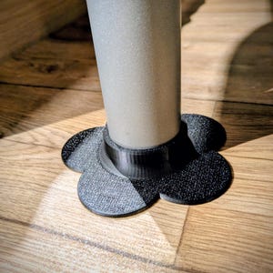 Puede incluir: Un protector de suelo para patas de muebles en forma de flor negra con superficie texturizada. La abertura circular central se ajusta a una pata de mueble cilíndrica de color gris claro, mientras que los seis pétalos redondeados se extienden.