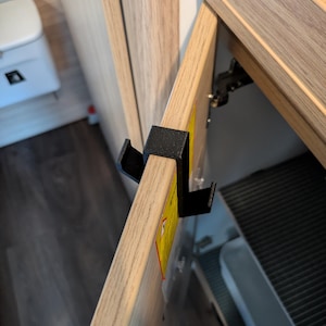 Peut inclure: Un crochet en plastique noir en forme de U monté sur le bord supérieur d'une porte de placard au fini bois clair. Le crochet présente une surface texturée et deux ergots orientés vers le haut sur le côté extérieur pour suspendre des objets.