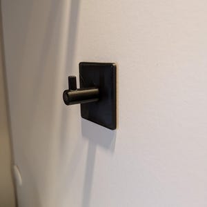Op de afbeelding: Een zwarte wandhaak met een vierkante basisplaat en een cilindervormige arm met een klein lipje aan het uiteinde dat naar boven wijst. De haak heeft een getextureerde afwerking en is bevestigd aan een vlak oppervlak voor het ophangen.