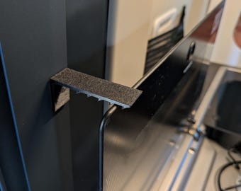 May include: A black, L-shaped bracket with a textured surface, mounted to a vertical black panel. The bracket features a series of small, downward-pointing teeth along its horizontal edge, which supports a thin, black, rectangular glass panel.