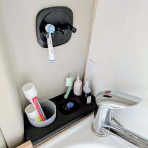 Puede incluir: Un soporte de pared negro para cepillos de dientes con un cabezal eléctrico colocado, y una bandeja organizadora negra sobre la encimera que contiene un tubo de pasta de dientes en un vaso, un cepillo manual y dos mangos eléctricos.