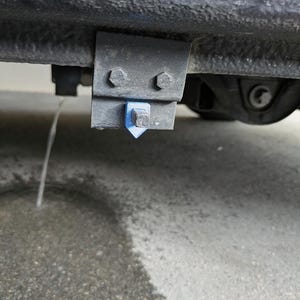 Op de afbeelding: Een zwarte metalen beugel gemonteerd aan de onderkant van een voertuigchassis, voorzien van twee zeskantbouten en een klein, felblauw, driehoekig plastic onderdeel in het midden, met een dunne straal vloeistof die naar beneden druppelt.