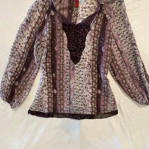 May include: A long-sleeved blouse featuring a vertical striped pattern in shades of purple, brown, and white. The garment has a deep V-neckline with a dark purple velvet-textured panel at the center and matching velvet trim along the bottom hem.