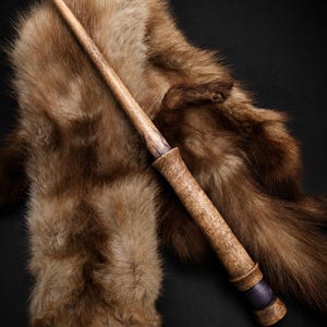 Peut inclure: Une baguette en bois fine avec un fût effilé brun clair au grain visible. La section de la poignée est plus épaisse, texturée avec un motif de bois naturel, et se termine par un anneau décoratif cylindrique violet foncé et une base évasée.