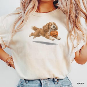May include: An ivory t-shirt featuring a graphic illustration of a brown spaniel dog running while carrying an orange jack-o'-lantern pumpkin bucket in its mouth. The dog has long, wavy fur and is depicted in a mid-air motion with a shadow below.