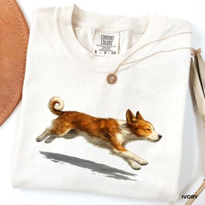 May include: An ivory-colored t-shirt featuring a graphic of a running brown and white dog. The shirt has a crew neckline and a Comfort Colors brand label at the inner collar. A thin gold-toned chain necklace with a small circular pendant rests on the fabric.
