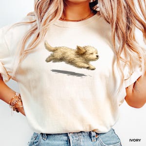 May include: An ivory-colored short-sleeved t-shirt featuring a graphic illustration of a small, fluffy, light-brown puppy in mid-air, appearing to leap or run. The puppy has closed eyes and a soft, textured coat, with a dark shadow beneath it.