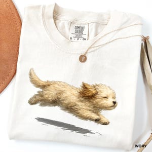 May include: An ivory t-shirt featuring a graphic of a running light brown puppy with fluffy fur. The shirt has a Comfort Colors label at the neck, size S, and is made of 100% cotton. A thin gold-toned chain necklace with a small round pendant rests on top.