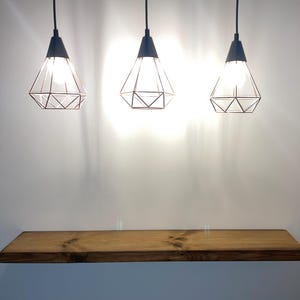 Op de afbeelding: Een rechthoekige zwevende houten plank met een natuurlijke houtnerfafwerking, gemonteerd aan een muur onder drie geometrische hanglampen van zwart metaal. Elke lamp heeft een ruitvormig draadframe en hangt aan een zwart snoer.