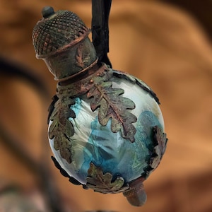 Op de afbeelding: Een bolvormig glazen ornament met een blauw en wit gemarmerd interieur, gevat in een donker, verweerd metalen frame in de vorm van eikenbladeren. De bovenkant is afgewerkt met een textuurrijk, eikelvormig metalen dopje in een donkere kleur.