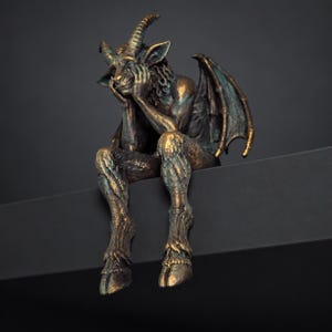 Puede incluir: Una figura de tono bronce de una criatura alada con cuernos sentada, apoyando la barbilla en sus manos. La escultura presenta pelaje texturizado, pezuñas hendidas y un acabado de pátina desgastada, en una pose contemplativa con las piernas colgando.