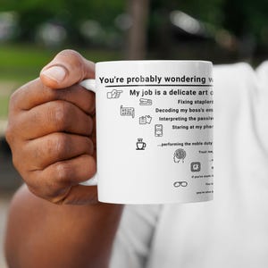 May include: A white ceramic mug featuring black text and icons. The text reads: You're probably wondering w... My job is a delicate art o... Fixing staplers, Decoding my boss's em..., Interpreting the passive..., Staring at my pho..., ...performing the noble duty, Trust me, ...a, B, If you've made it, You h..., you're also s.