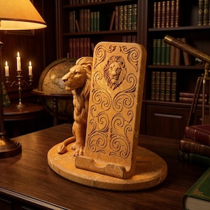May include: A tan stone phone stand featuring a sculpted lion figure sitting beside a rectangular upright panel. The panel displays an embossed lion head at the top and intricate swirling scrollwork patterns across its surface.