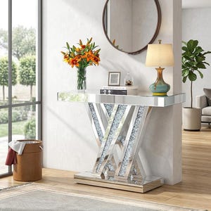 May include: A mirrored console table featuring a V-shaped base with inlaid crushed crystal accents. The rectangular top surface holds a glass vase with orange lilies, a small picture frame, a stack of books, and a table lamp with a beige shade.