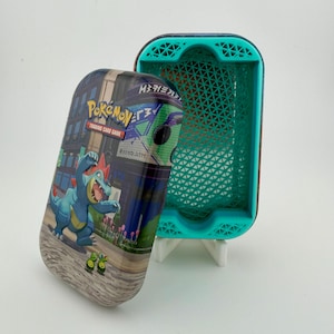 May include: A rectangular metal Pokemon Trading Card Game tin with a detached lid featuring a blue Feraligatr character. The interior of the tin is lined with a bright teal, 3D-printed plastic insert that has a geometric, triangular grid pattern.