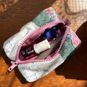 May include: A rectangular patchwork zipped pouch featuring various floral and paisley fabric patterns in shades of green, pink, and white. The pouch is open, revealing a small blue jar, a clear bottle with a white cap, and a grey cylindrical object.