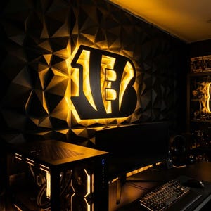 May include: A wall-mounted Cincinnati Bengals logo sign featuring a black cutout letter B with a vibrant orange LED backlight, positioned against a dark, geometric, three-dimensional textured wall panel surface in a gaming room setup.