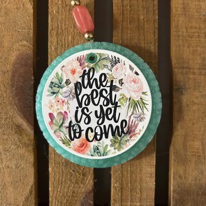 May include: A round hanging ornament with a teal textured border and a central white disc featuring a floral design of succulents and flowers. The text "the best is yet to come" is printed in black script. A pink bead hangs from the top string.