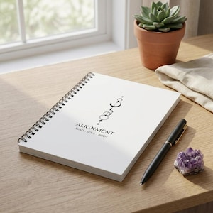 May include: A white spiral-bound notebook featuring a black graphic of celestial symbols and the text ALIGNMENT MIND - SOUL - BODY. The notebook is positioned next to a black ballpoint pen and a small purple amethyst crystal cluster.
