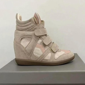 May include: A high-top wedge sneaker in shades of beige and soft pink. The shoe features a suede upper with multiple hook-and-loop velcro straps across the front, a padded collar, and a solid brown rubber sole. The design includes cut-out side panels.